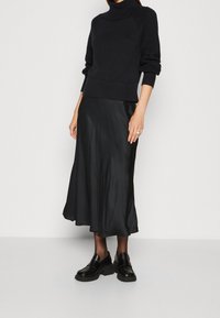 Schwarzer gestrickter Rollkragenpullover mit strukturierten Ärmeln, kombiniert mit einem fließenden schwarzen Satin-Midi-Rock und klobigen schwarzen Slippern.