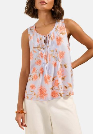 Frau trägt eine ärmellose weiße Bluse mit rosa Blumenmuster und einer Schleife am Ausschnitt, kombiniert mit weißen Hosen und einem Armband.