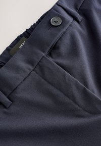 Un pantalon en tissu bleu marine présente une texture lisse, une fermeture à boutons et une taille élastiquée avec des poches latérales pour une utilité supplémentaire.