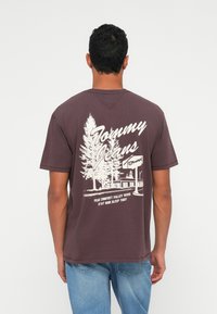 Bordo puuvillane t-särk, mille seljal on valge graafika puude ja maja kujutisega, tekstiga: "Tommy Jeans" ja selle all olev loosung.