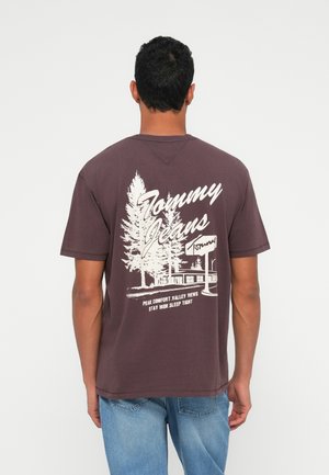 Bordeaux farvet bomulds t-shirt med en hvid grafisk illustration af træer og en hytte på bagsiden, med teksten: "Tommy Jeans" og slogan nedenunder.