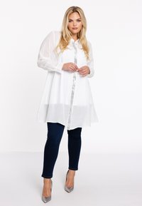 Witte blouse met lange mouwen, knoopsluiting en transparante stof, gecombineerd met donkerblauwe skinny jeans en zilveren hoge hakken.