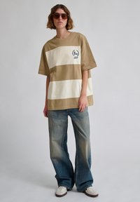Beige og creme stribet oversized t-shirt med rund halsudskæring og broderede detaljer på venstre bryst, parret med brede blege blå jeans.