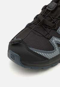Chaussure de sport noire et grise avec un tissu texturé, un capuchon en caoutchouc pour les orteils, des lacets élastiques et un motif rayé sur les côtés.