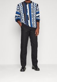 Pull bleu à motifs multiples avec manches longues et poignets côtelés, associé à un pantalon noir. Le modèle porte des chaussures noires.