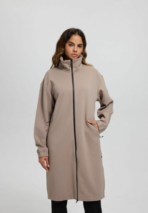 Donna con capelli lunghi e mossi che indossa un cappotto color taupe, lungo fino al ginocchio, con chiusura a zip, collo alto e tasche laterali, in piedi davanti a uno sfondo neutro.