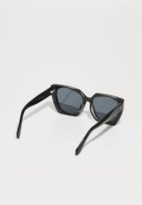 Occhiali da sole in acetato nero con una montatura geometrica, lenti grigio scuro e finitura lucida. Presenta aste nere sottili con sottili dettagli dorati.