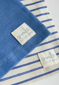 Vêtements pour bébé à rayures bleues et blanches et unis bleus avec étiquettes cousues portant l'inscription « souris petit » sur une surface claire.