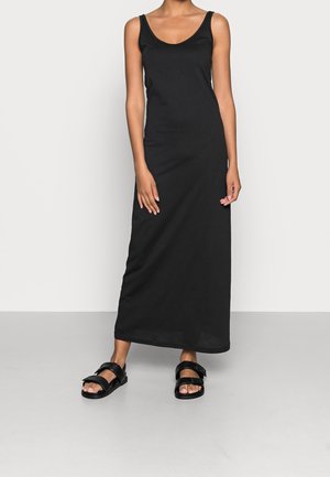 Frau trägt ein ärmelloses schwarzes Maxikleid und schwarze Sandalen mit offenen Zehen, steht vor einem schlichten hellen Hintergrund.