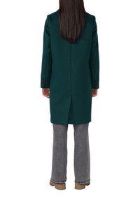Donkergroen wollen jas met een rechte snit, enkelventilatie en knoopmanchetten, gecombineerd met grijze broek en beige schoenen.