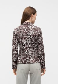 Γυναίκα που φοράει εφαρμοστό σακάκι με λεοπάρ σχέδιο και μακριά μανίκια, σε συνδυασμό με ανοιχτό γκρι τζιν, όρθια και γυρισμένη πλάτη, σε απλό φόντο.