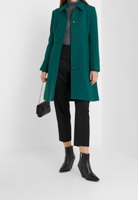 Teal lange jas met een kraag en knoopsluiting, gecombineerd met een grijze coltrui, zwarte cropped broek en zwarte enkellaarsjes.