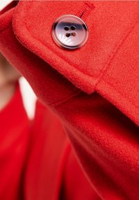 Abrigo de lana rojo con un acabado texturizado, con un botón de plástico transparente de cuatro agujeros. Primer plano de la zona del hombro y detalle del botón.