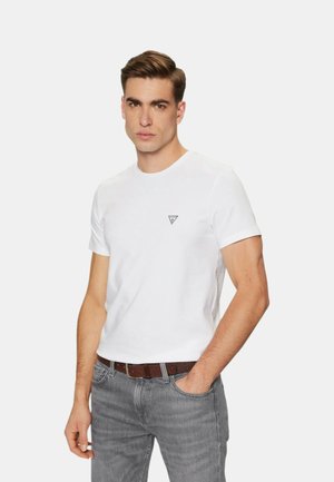 Uomo con capelli castani corti che indossa una maglietta bianca aderente con un piccolo logo e jeans grigi, posando con una mano in tasca su sfondo bianco.