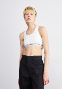 Soutien-gorge de sport blanc à encolure dégagée et bande grise avec logo. Pantalon noir taille haute avec poches latérales et détails de couture subtils.