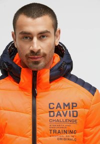 Orange padded jacket with navy accents, high collar, and zip closure. Features logo and text print on the chest. Quilted texture visible.