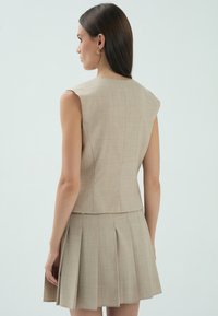 Beżowa, dopasowana bezrękawnik z dopasowanym korpusem, w połączeniu z plisowaną mini spódnicą, wykonaną z teksturowanej tkaniny.