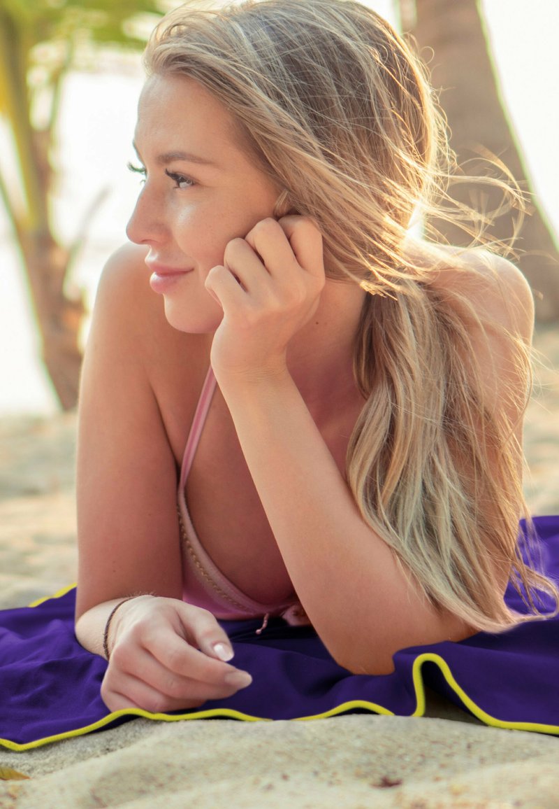 Eine Person mit langen blonden Haaren liegt auf einer Sandfläche, trägt ein rosa Oberteil und ist von einem violetten Stoffhandtuch mit gelbem Rand umgeben.