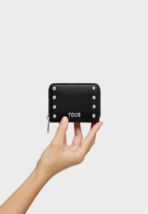 Mano sosteniendo una cartera de cuero negro con cremallera, tachuelas plateadas y el logo de TOUS sobre un fondo liso gris claro.