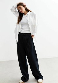 Weißes Button-up-Hemd über einem weißen, figurbetonten Oberteil, kombiniert mit lockeren schwarzen Hosen. Die Schuhe haben eine cremefarbene und tannfarbene Farbgebung. Glattes Material, lockere Passform.