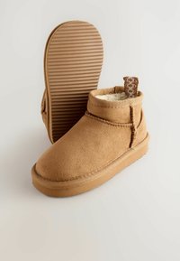 Bottes de cheville en daim tan avec une doublure en polaire, semelle en caoutchouc rainurée plate et une boucle tissée à l'arrière pour un enfilage facile. Conception à bout arrondi.