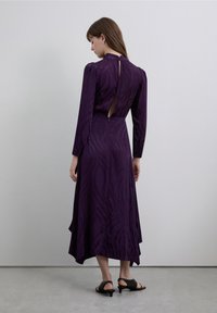 Vestido midi morado con mangas largas, detalle de abertura en la espalda y dobladillo fluido. Confeccionado con tela suave que presenta un patrón sutil. Sandalias negras de tacón.