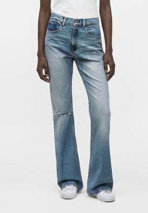 Personne portant un jean bleu clair délavé avec une petite déchirure au genou, des baskets blanches et un haut blanc rentré, debout devant un fond uni.
