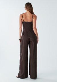 Žena v bezramenném, širokém tmavohnědém overal s náramky, stojící zády na bílém pozadí.