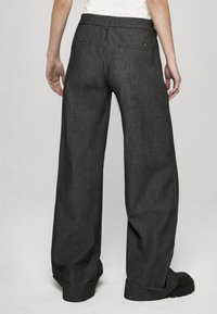 Kalhoty se širokými nohavicemi v tmavě šedé tkanině s jemnou texturou, zadními kapsami a ohrnutými lemy. Černá obuv doplňuje vzhled.