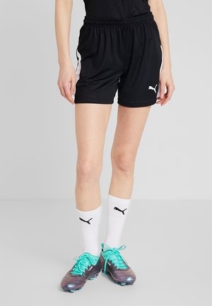 Person trägt schwarze Sportshorts mit weißem Puma-Logo und Streifen, weiße Puma-Socken und lila Fußballschuhe mit türkisfarbenen Schnürsenkeln.