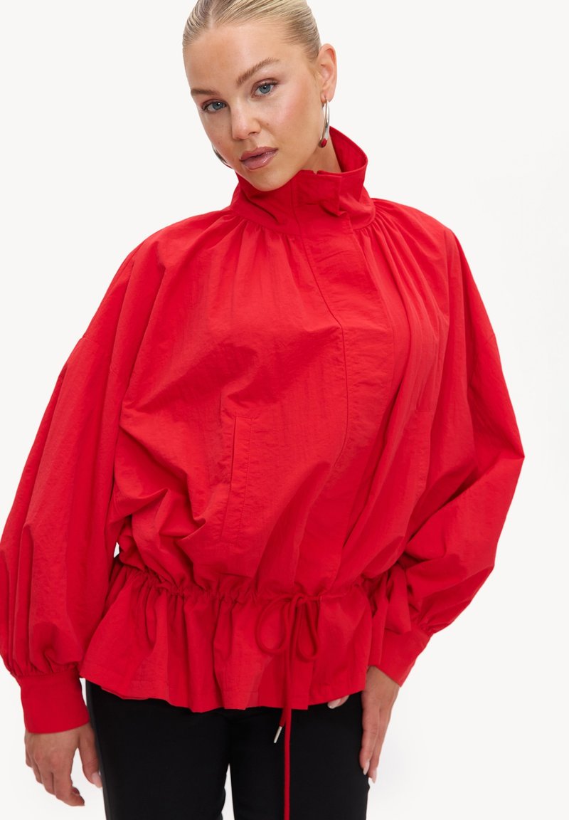 Femme aux cheveux plaqués en arrière porte une veste rouge vif, ample avec un col montant et une taille cintrée, associée à un pantalon noir.