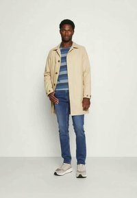 Trench coat bege com botões, camisa riscada azul, jeans azuis e ténis cinzentos. Tecido suave, ajuste definido e estilo casual.