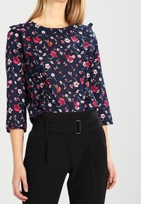 Kobieta w granatowej bluzce w kwiaty z falbanami i czarnych spódnicach z wysokim stanem z paskiem, stojąca na tle jednolitej ściany.