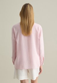 Pink long-sleeve shirt with a textured fabric, featuring a relaxed fit and button detailing on the sleeves. Paired with white shorts.