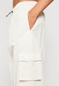 Bílé cargo kalhoty Nike, lehký materiál, elastický pas, boční kapsa s klopou, viditelné logo, texturovaný materiál.