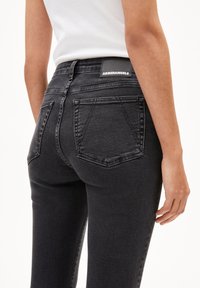 Hoge taille zwarte denim jeans met een slank ontwerp, voorzien van achterzakken en een leren patch met het label "ARMEDANGELS" op de taille.