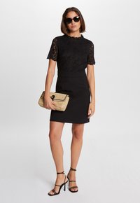 Zwarte kanten top met korte mouwen, gecombineerd met een aansluitende zwarte rok. Accessoires zijn een geweven clutch en zwarte bandjeshakken.