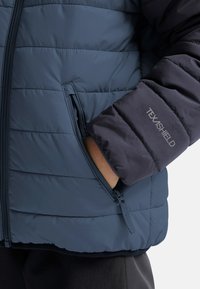 Navy puffjacka med quiltat mönster, utrustad med en blixtlåsficka och "TEXSHIELD"-logotyp på ärmen, tillverkad av lätt material.