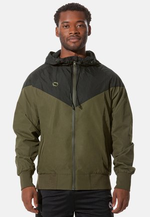 Olivegrüne Jacke mit schwarzer Einfassung, Reißverschluss vorne, verstellbare Kapuze, elastische Bündchen und einem Logo auf der oberen linken Seite.