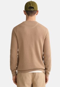Pull en tricot beige avec un motif texturé, col rond, poignets et ourlet côtelés. Porté sur une chemise blanche, associé à un pantalon beige clair.