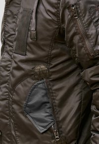 Närbild av en brun jackärm som visar en vertikal dragkedjeficka och sydda detaljer, vilket framhäver textur och designelement.