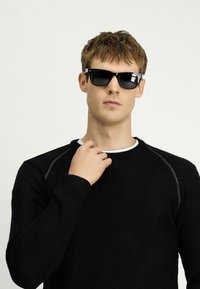 Czarne okulary przeciwsłoneczne o prostokątnym kształcie. Noszone przez osobę w czarnej swetrze o fakturze, z prążkowanym dekoltem i mankietami.