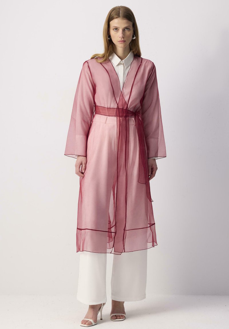 Chiuso kimono trasparente di colore rosa con cintura color marrone scuro, abbinato a pantaloni bianchi a gamba larga e tacchi chiari; presenta maniche lunghe e dettagli delicati.