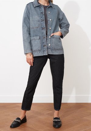 Persoon draagt een lichtblauw denimjack met knopen, zwart t-shirt, zwarte cropped jeans en zwarte loafers met zilveren kettinkdetail.