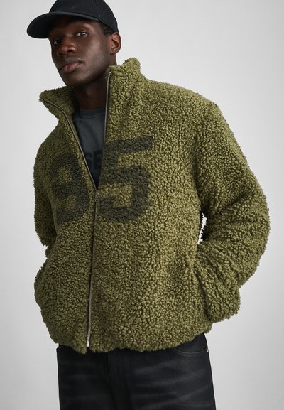 Veste en polaire vert olive avec une surface texturée, col montant, fermeture éclair et un grand '95' imprimé en noir sur le devant.