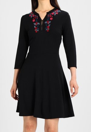 Robe noire longueur genou avec manches trois-quarts et broderie florale rouge et bleue autour de l'encolure, avec un détail de fermeture éclair.