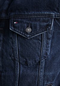 Detalle de chaqueta de mezclilla azul oscuro con un bolsillo en el pecho con un botón negro y una etiqueta de logotipo en rojo, blanco y azul. Tela texturizada y costuras visibles.