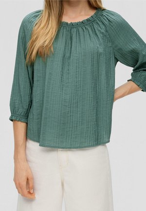 Žena nosící zelenou texturovanou halenku s dlouhým rukávem a nabíraným výstřihem a krémové vysokým pasem kalhoty, pózující s jednou rukou na bok.