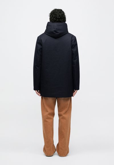 Meresinine pikk mantel kapuutsiga, sirge lõike ja sileda tekstuuriga, kombineeritud helepruuni laiade pükstega.