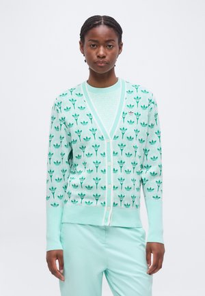 Nő világos türkiz kardigánban és nadrágban, melyeken ismétlődő sötét türkiz Adidas trefoil logók vannak, egyszerű fehér háttér előtt áll.